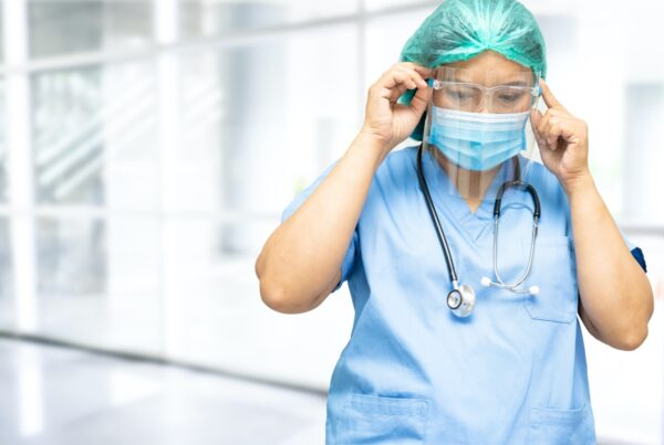 Healthcare worker adjusting face shield and goggles while wearing a surgical mask, cap, and stethoscope.