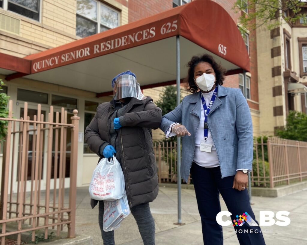 Woman in face shield holding a bag of PPE bumps elbows with a woman in a blazer and a white face mask under a red awning that reads 'Quincy Senior Residences.'